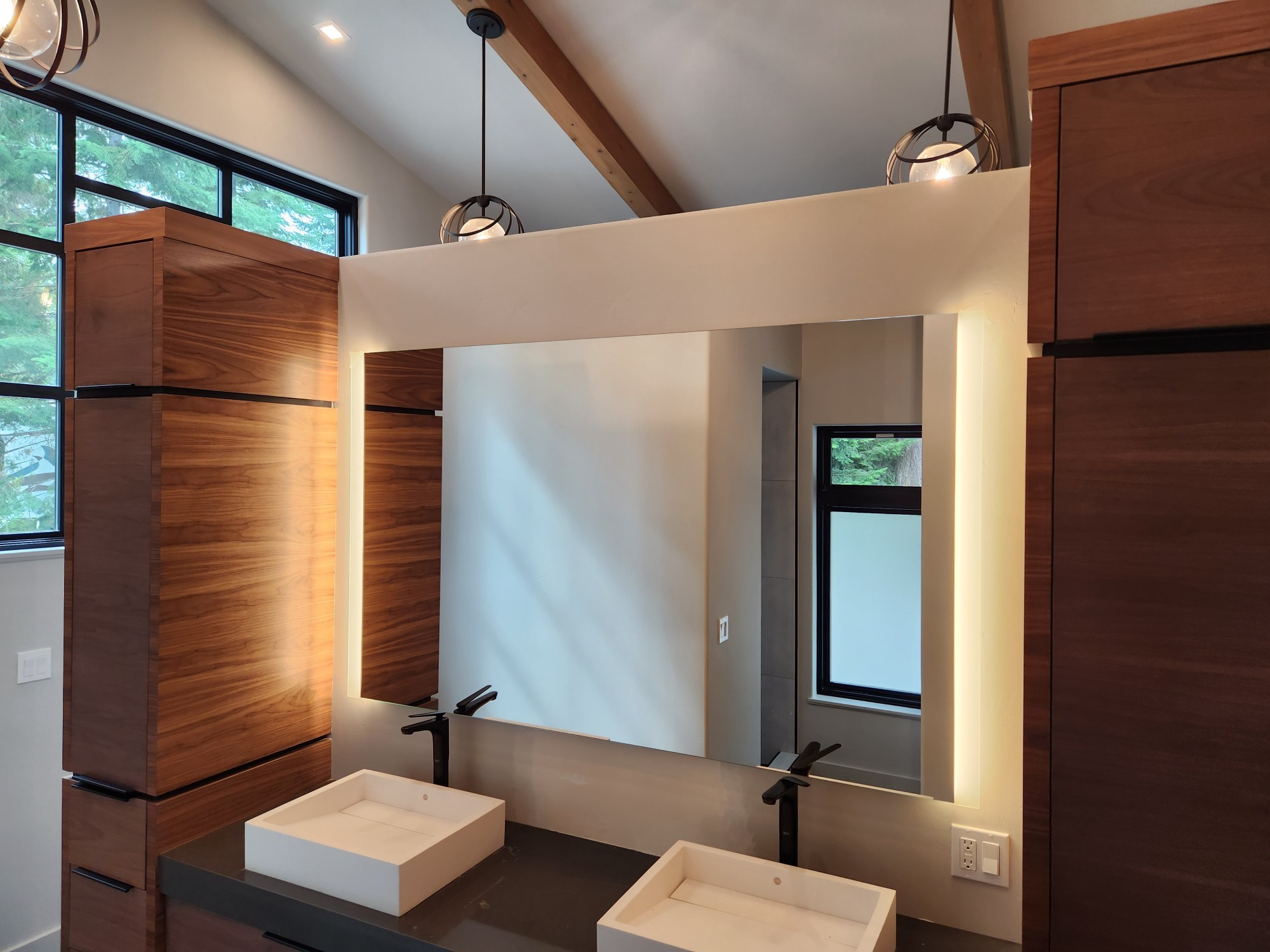 A bathroom with tall wood-grain cabinets, two square vessel sinks with black faucets on a dark counter, a large backlit mirror, pendant lights and an exposed timber beam