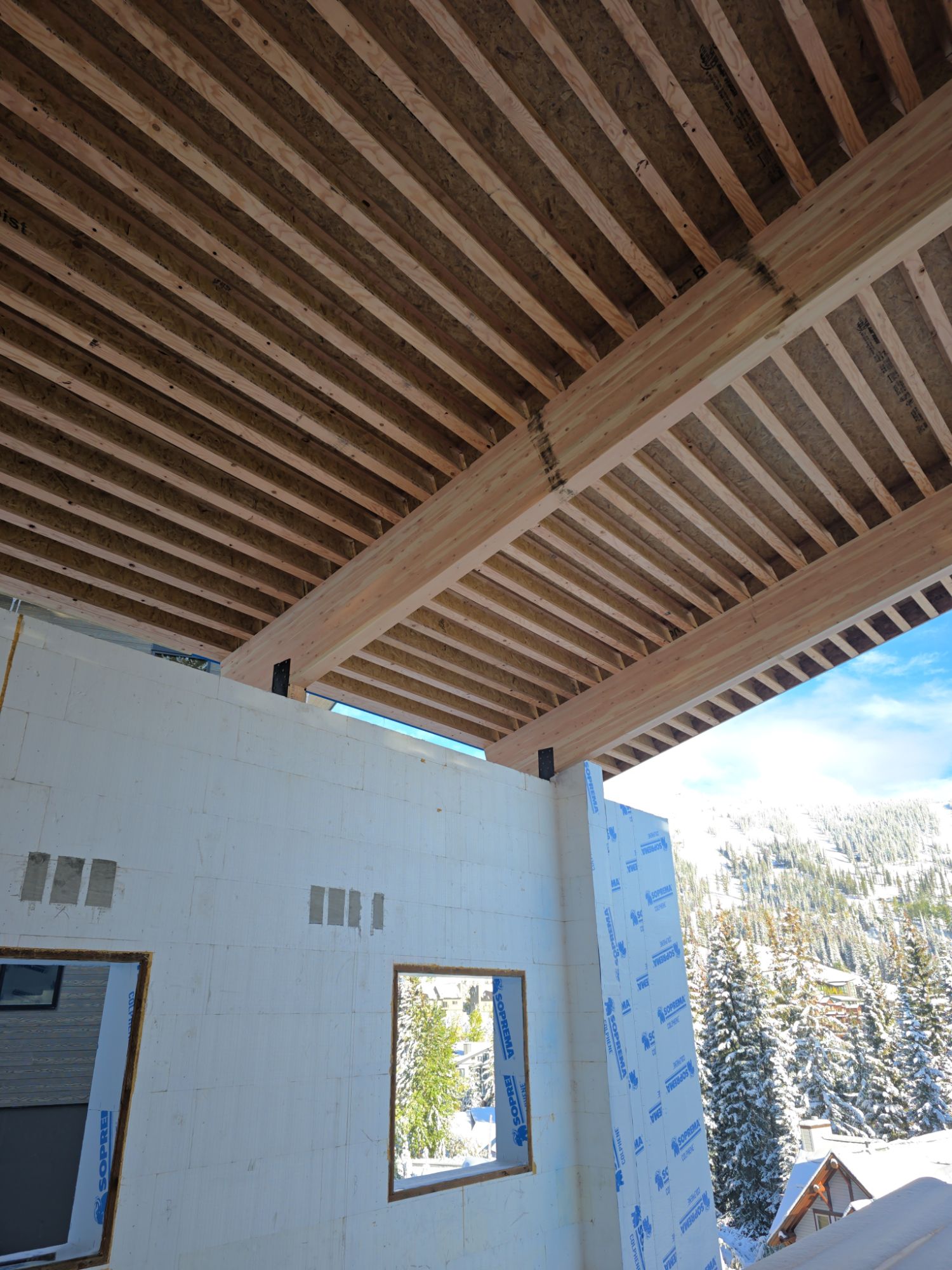 Heavy timber beams and open roof joists over a home under construction, its walls wrapped in white membrane, with snowy evergreens and a ski hill beyond