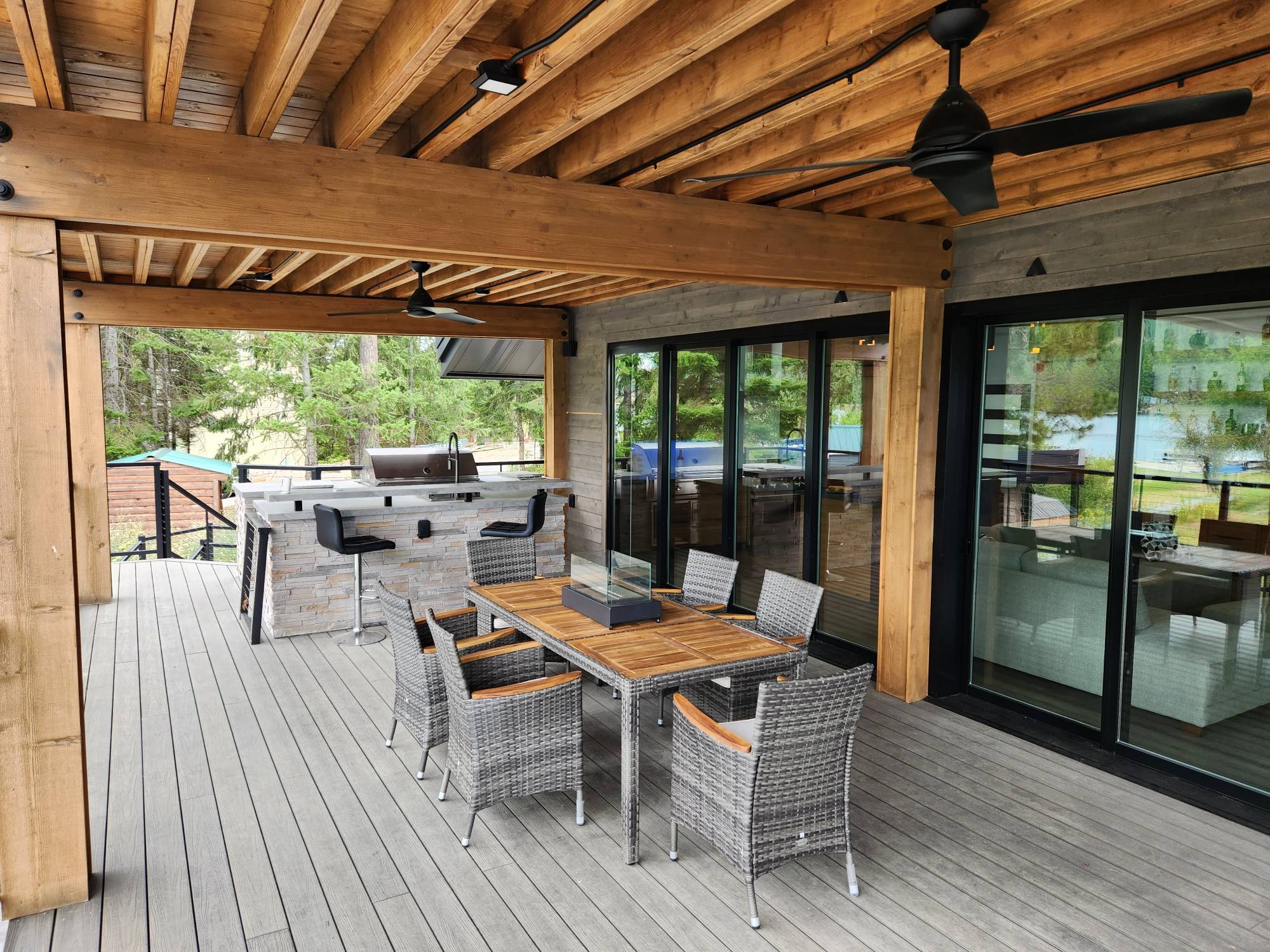A covered deck under exposed timber beams and ceiling fans, with a stacked-stone outdoor kitchen and bar, a dining table with wicker chairs, and sliding glass doors into the house