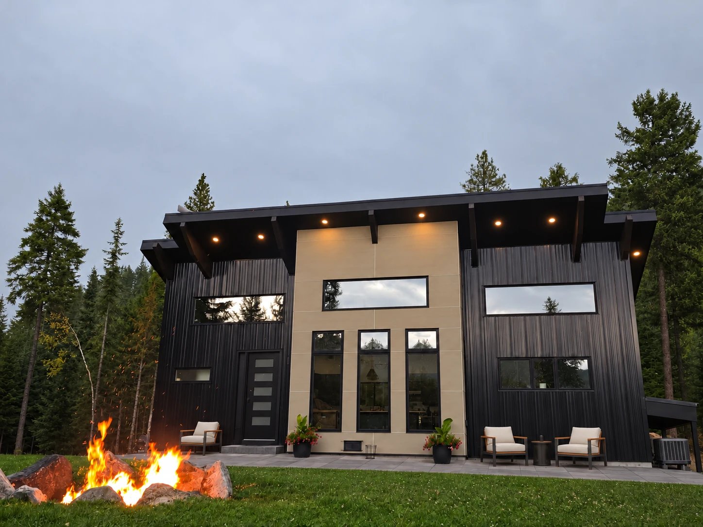 A two-story modern home with black ribbed siding, a tan center section with tall black-framed windows and a deep lit overhang, seen across a lawn with a stone fire pit, among evergreens