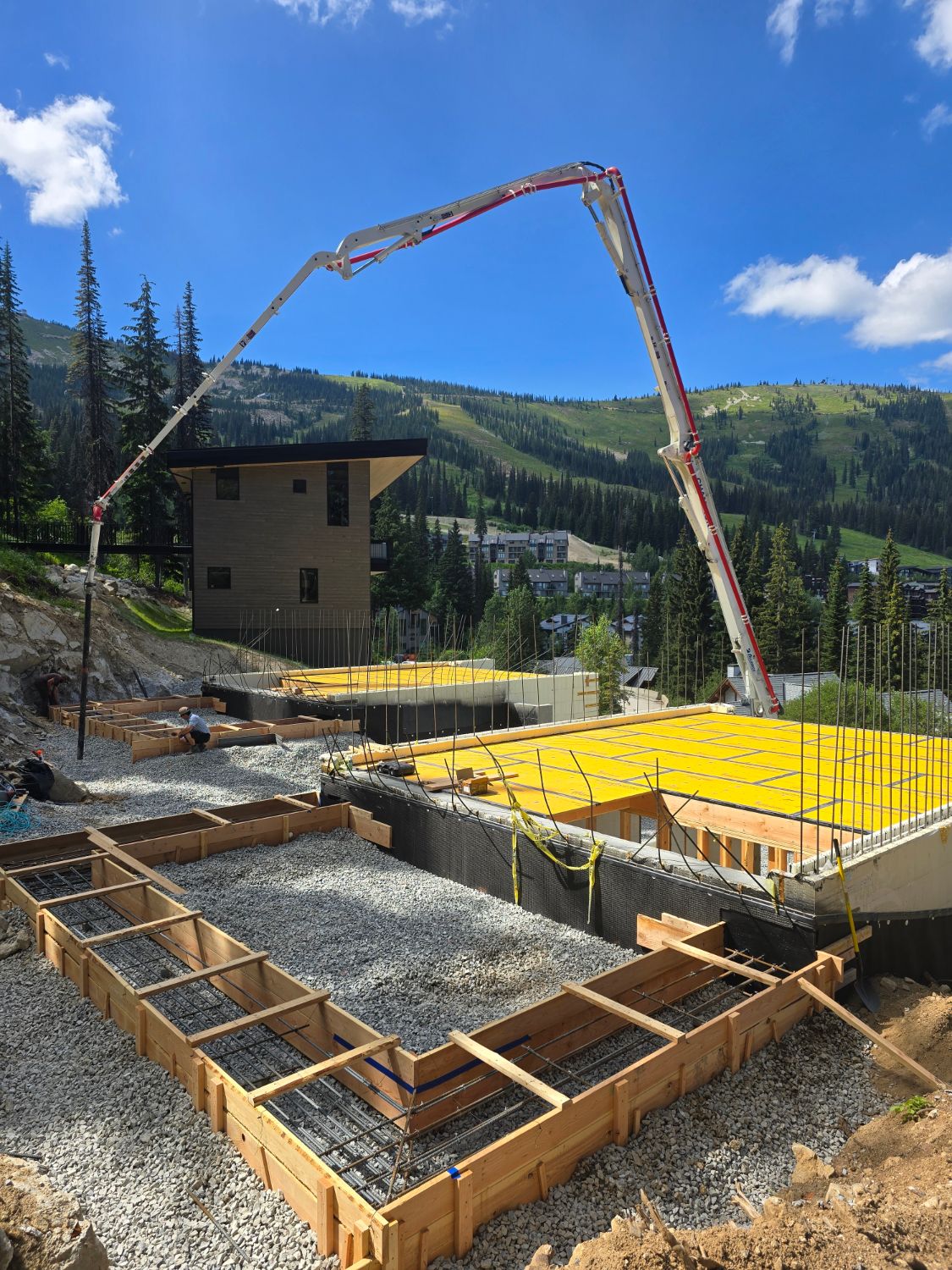 A concrete pump boom arching over a hillside foundation, with rebar-filled footing forms in the foreground, a finished modern house beside it and a ski hill behind