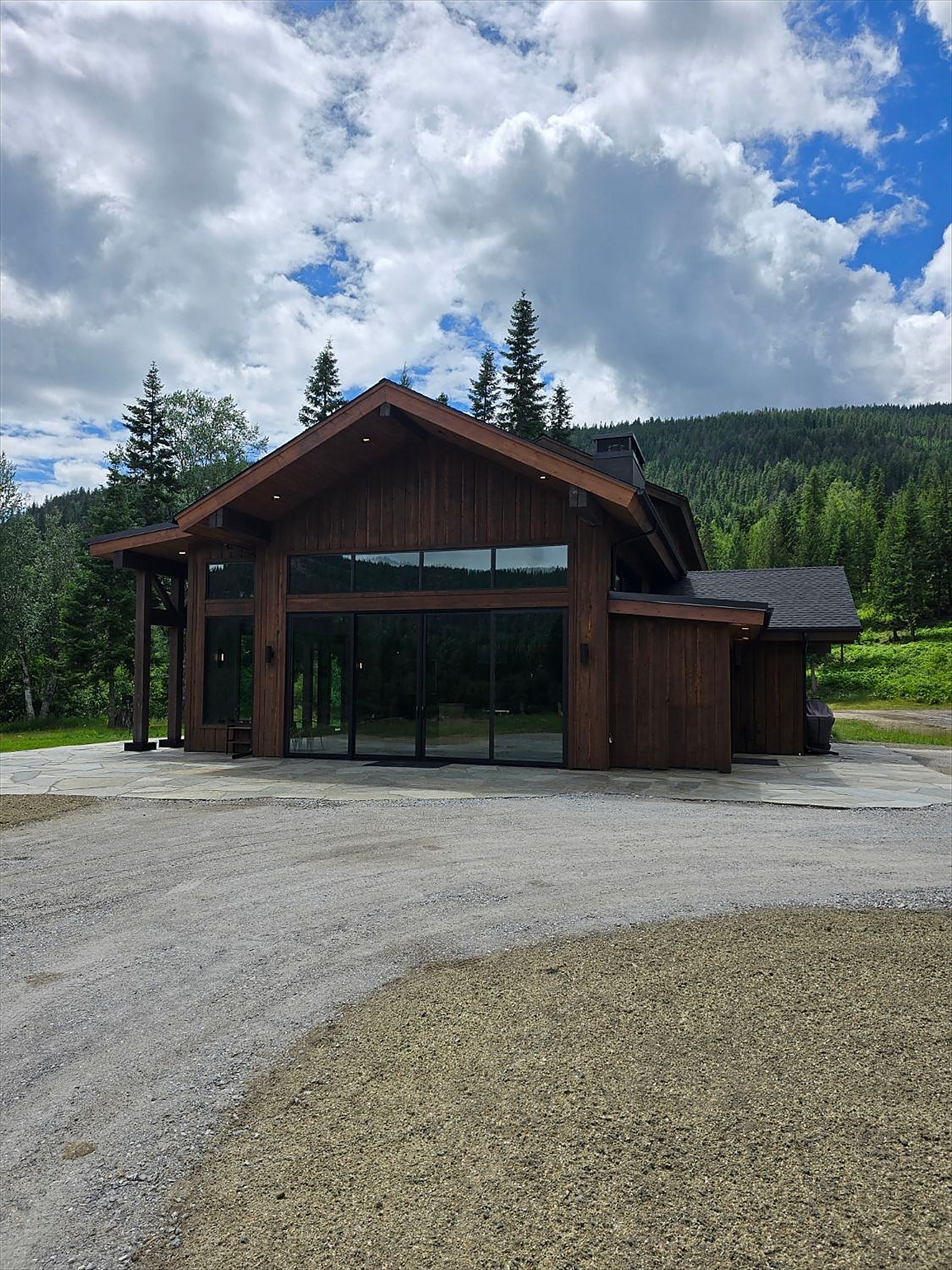 A home with dark-stained board siding, a gabled roof and a full-height wall of glass, on a stone patio below forested hills